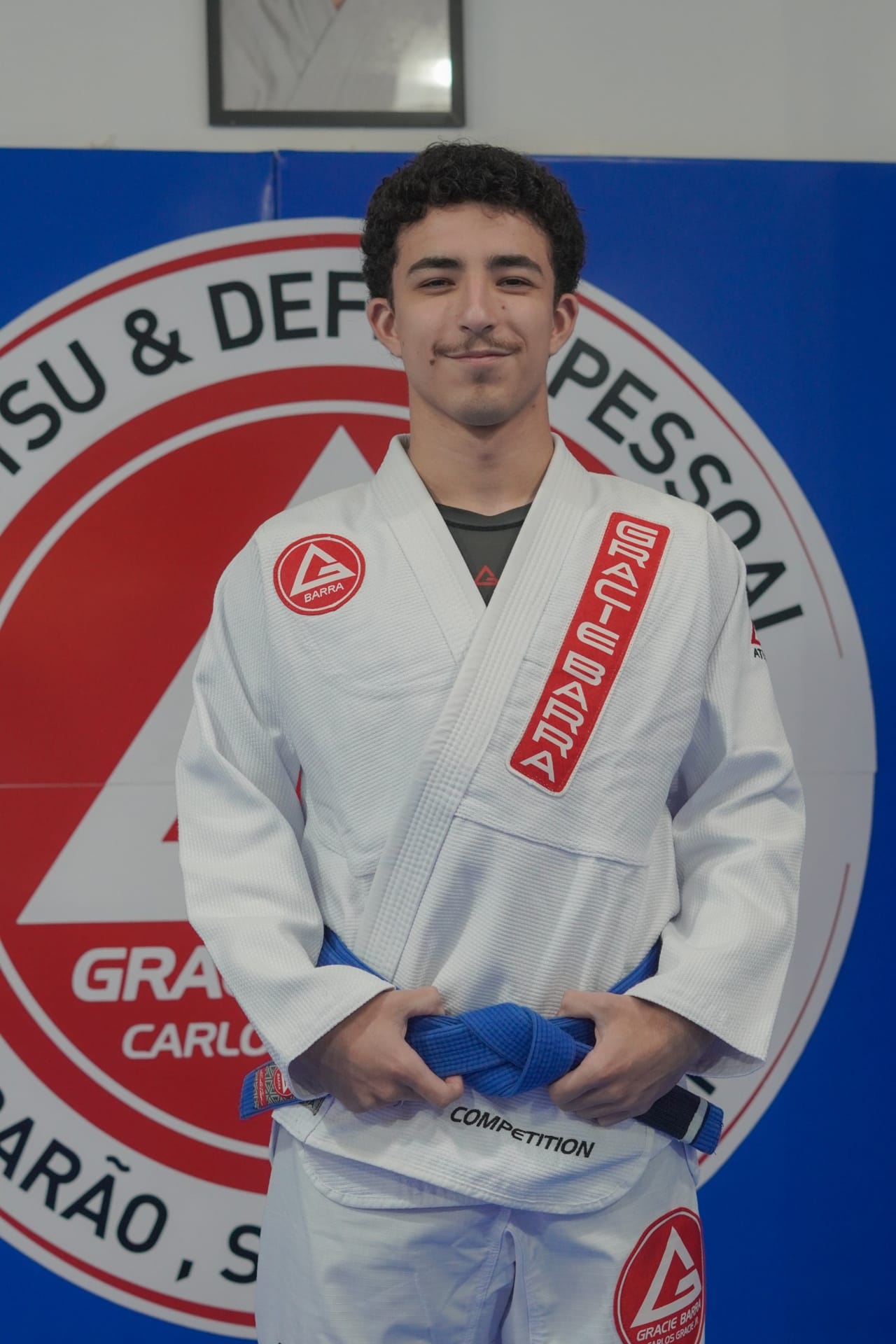 Alysson, faixa azul, sorrindo diante do escudo da GB Tubarão