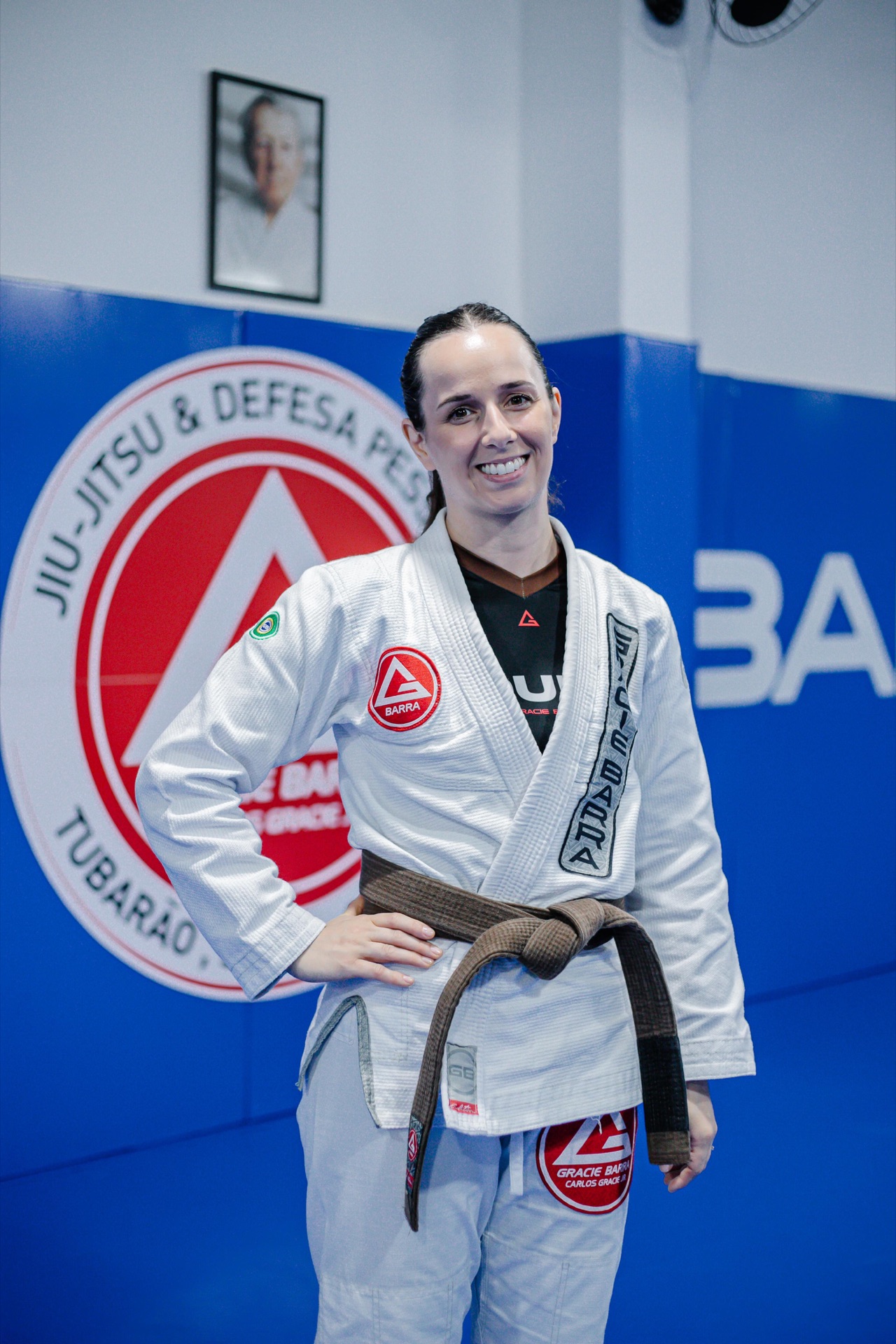 Instrutora Glaucia, faixa marrom, sorrindo diante do escudo da GB Tubarão