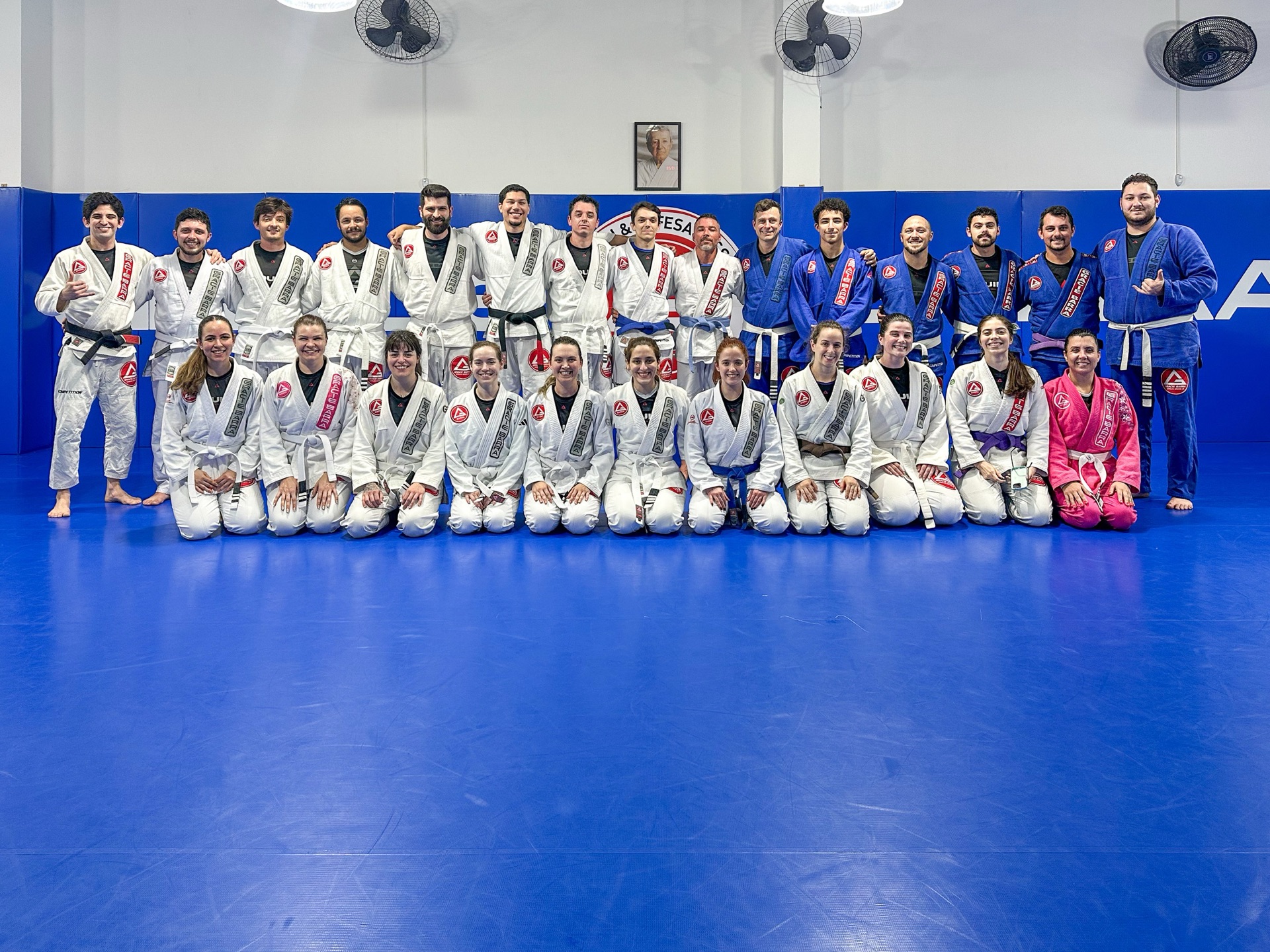 Turma adulta mista da Gracie Barra Tubarão reunida no tatame azul, alunos de várias faixas sentados e em pé